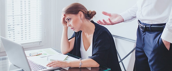 An unhappy employee sitting at a laptop looking upset with a man standing behind her.